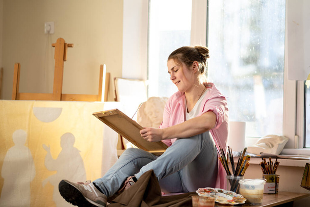 Fehér nő művész, aki egy festményen dolgozik fényes nappali stúdióban. Happy művész rajzol egy művészeti projekt festékek és ecsettel a műhelyben. Hobbi. Művész a munkahelyemen. Kreatív szakma. - Fotó, kép