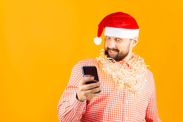 een man in een rood geruit hemd, een kerstman hoed en een bloemenslinger om zijn nek houdt een smartphone voor zich - Foto, afbeelding