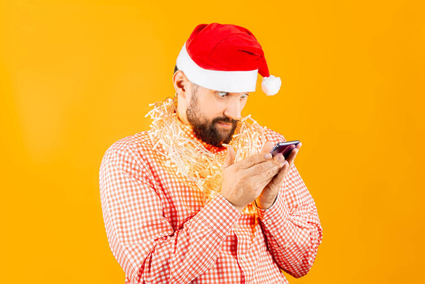 Sarı arka planda, kırmızı ekose gömlekli ve Noel Baba şapkalı bir adam önünde iki eliyle akıllı telefonu tutuyor. - Fotoğraf, Görsel