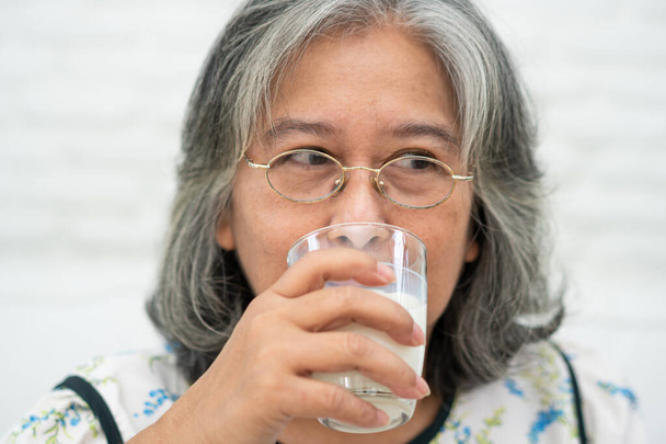 Asian Senior woman holding glass milk while relaxing on a sofa living room for retirement wellness. Elderly Woman drinking a glass of milk to maintain her wellbeing. Concept of wellness and healthy. - Photo, Image