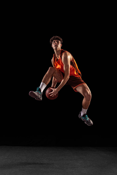 Ganzes Porträt des Basketballtrainings isoliert auf dunklem Studiohintergrund. Großer muskulöser Athlet springt mit Ball. - Foto, Bild