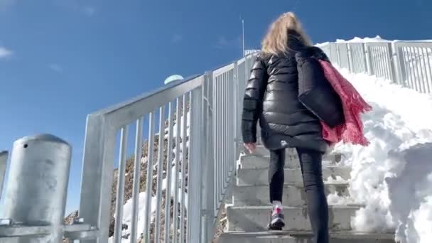 Взрослая женщина в пальто, солнцезащитных очках и маске для лица, поднимаясь по лестнице, с радостью простирает руки на Леднике 3000. Концепция туризма и путешествия. - Кадры, видео