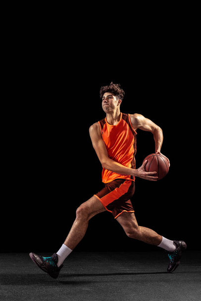 Full length portrait of basketball player training isolated on dark studio background. Tall muscular athlete jumping with ball. - Foto, Imagen