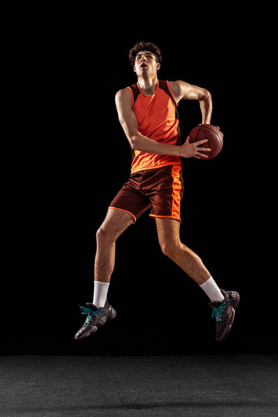 Full length portrait of basketball player training isolated on dark studio background. Tall muscular athlete jumping with ball. - Фото, зображення