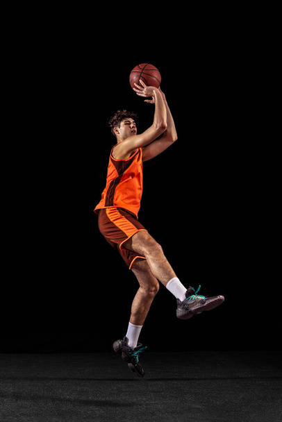 Full length portrait of basketball player training isolated on dark studio background. Tall muscular athlete jumping with ball. - Photo, Image