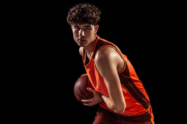 Close-up portrait of basketball player posing with ball isolated on dark studio background. Sport, energy, power, results. - Φωτογραφία, εικόνα