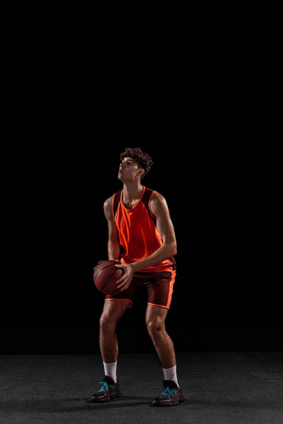 Full length portrait of basketball player training isolated on dark studio background. Tall muscular athlete jumping with ball. - Fotó, kép