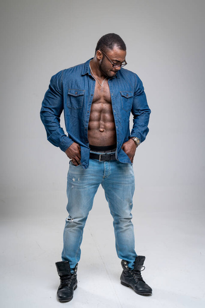 Handsome African guy with naked torso wearing jeans shirt. Athletic man in denim sanding on white background. Bodybuilder male model wearing glasses. - Foto, immagini