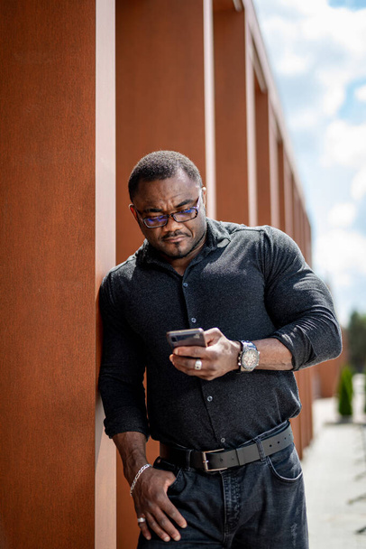 African man standing at the building and holding phone in his hand. Black man in dark clothes leaning on the wall of a building. Sportive guy in black checking up his mobile at business center. - Фото, изображение