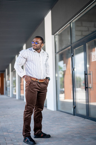 African man in white shirt and jeans standing at the doors of business center. Sportive male holding his hands in the pockets standing in the street. Businessman standing at the door of business center. - 写真・画像