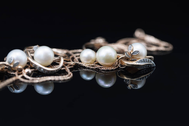 Goldene Ohrringe und Ringe mit weißen Perlen liegen auf einer schwarzen Spiegeloberfläche - Foto, Bild