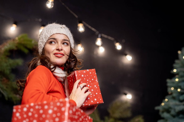 Beautiful lady in white winter cap holding Christmas presents in her hands. Happy girl under the decoration light garland near the Christmas tree. Smiling woman with gifts in the black background. - Foto, immagini