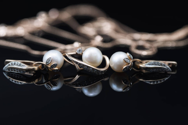 Goldene Ohrringe und Ringe mit weißen Perlen liegen auf einer schwarzen Spiegeloberfläche - Foto, Bild