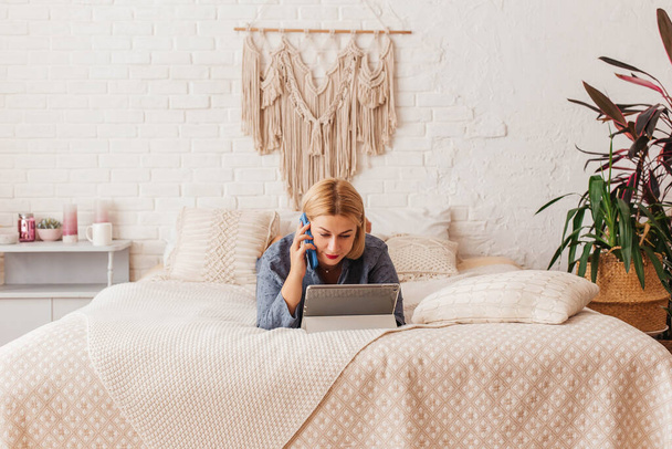 Mooie jonge vrouw in pyjama, werkend op een laptop in bed, pratend aan de telefoon. Online winkelen - Foto, afbeelding