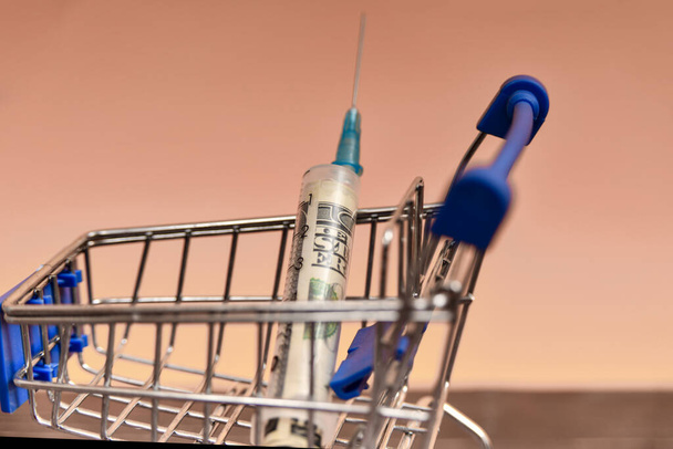 Injekční stříkačka s dolarem uvnitř leží v nákupním košíku. - Fotografie, Obrázek