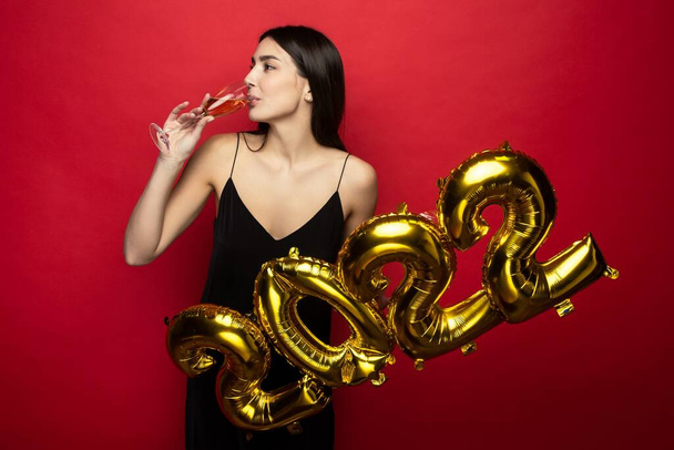 Eine junge schöne Frau im schwarzen Kleid hält die Zahlen 2022 und ein Glas Champagner in der Hand. - Foto, Bild