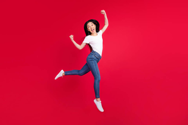 Photo of astonished crazy korean lady jump raise fists wear white t-shirt jeans sneakers isolated red color background - Photo, image