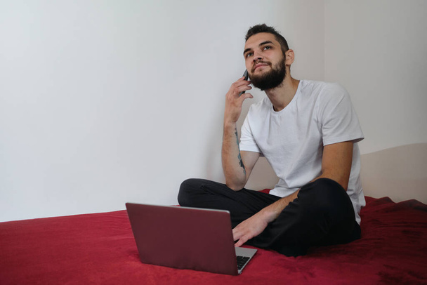 Young handsome freelancer man in white Tshirt with beard and dreadlocks works at home on self isolation. Conduct business via Internet online. Talk on phone and discuss important matters. - Photo, Image