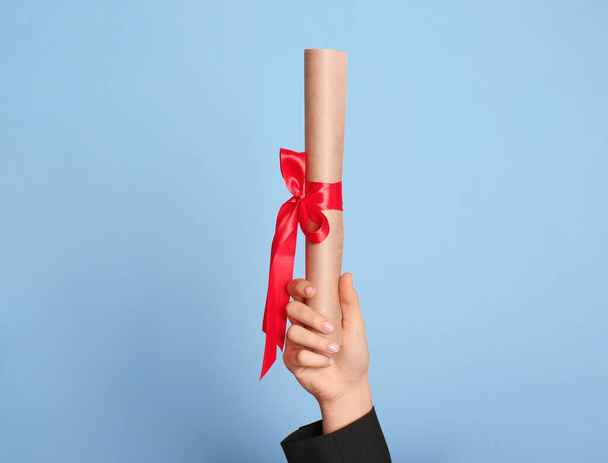 Student holding rolled diploma with red ribbon on light blue background, closeup - Foto, afbeelding