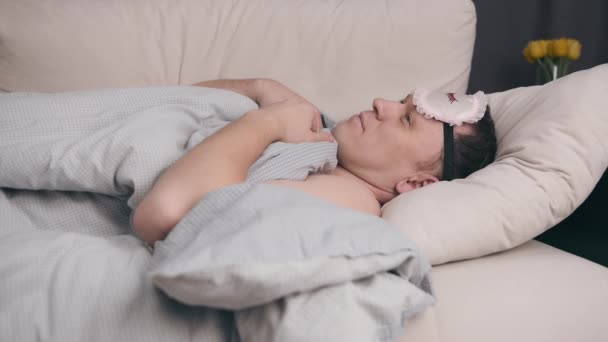 Uyku maskeli genç bir adam battaniyeyle kaplı kanepede yatıyor, gözlerine maske takıyor ve uykuya dalıyor. - Video, Çekim