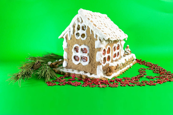 Házi készítésű mézeskalács ház festett fehér édes cukormáz és díszített fenyőággal zöld képernyős háttérrel. A finom, kézzel készített édességek nyaralásra való elkészítésének koncepciója. - Fotó, kép