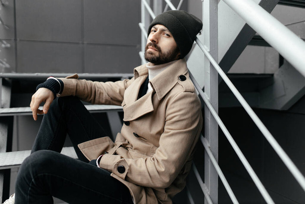 O moderno hipster urbano com barba e bigode está sentado em uma velha escadaria de ferro na cidade. Um cara atraente relaxa ao ar livre. - Foto, Imagem
