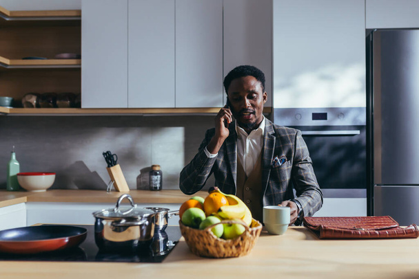 African American businessman going to work at home, having breakfast in the kitchen, talking on the phone - Foto, imagen