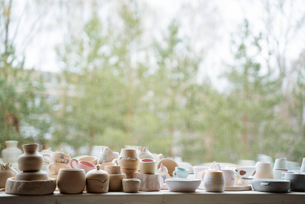 Many ceramics, mainly mugs and vases, stand on a bright windowsill in the workshop. Outside the window there are green trees. High quality photo - Photo, Image