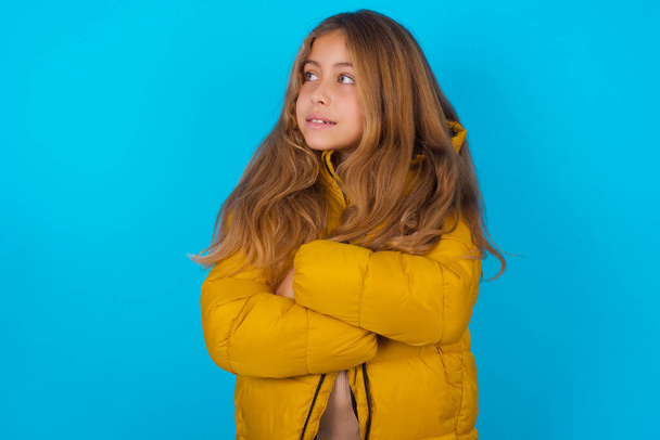 Dromerige rust ontspannen brunette kid meisje dragen gele jas over blauwe achtergrond kruising armen, ziet er goed copyspace - Foto, afbeelding