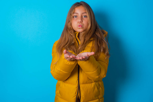 μελαχρινή κορίτσι παιδί φορώντας κίτρινο σακάκι πάνω από το μπλε φόντο στέλνοντας φιλί χτύπημα με στόμιο χείλη και κρατώντας παλάμες για να στείλετε φιλί αέρα. - Φωτογραφία, εικόνα