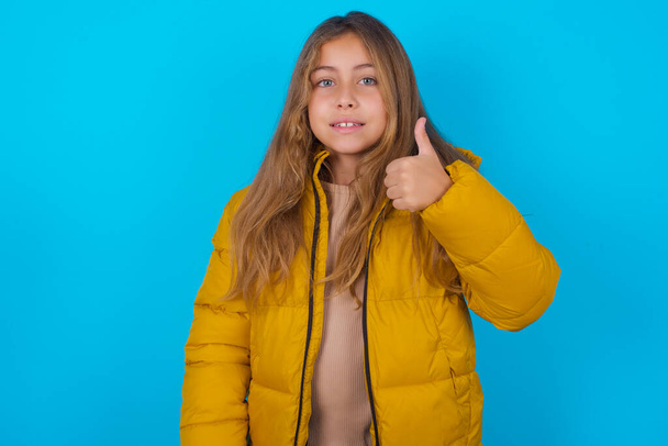 fille brune enfant portant une veste jaune sur fond bleu faisant des pouces heureux geste avec la main. Approuver l'expression en regardant la caméra montrant le succès. - Photo, image