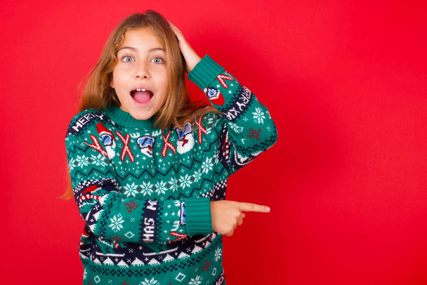 krásná dívka na sobě vánoční svetr pocit pozitivní má užaslý výraz, naznačuje něco. Jednou rukou na hlavě a druhou rukou ukazuje. - Fotografie, Obrázek