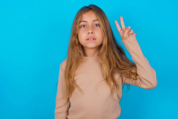 brunette kid meisje dragen trui over blauwe achtergrond tonen en wijzen met vingers nummer twee terwijl glimlachen zelfverzekerd en gelukkig. - Foto, afbeelding