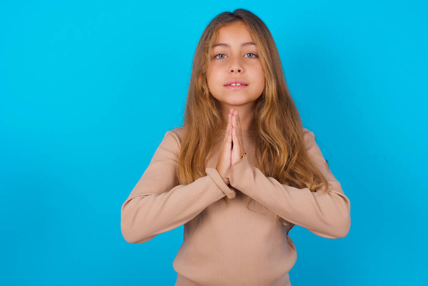 brunette kid meisje dragen trui over blauwe achtergrond bidden met de handen bij elkaar vragen om vergeving glimlachen zelfverzekerd. - Foto, afbeelding