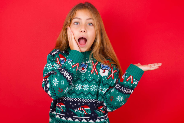 Positiivinen kaunis tyttö yllään joulu pusero tyttö sanoo: wow kuinka jännittävää se on, osoittaa jotain. Yksi käsi hänen päänsä päällä ja osoittaa toisella kädellä. - Valokuva, kuva