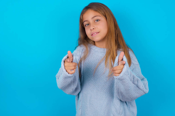barna gyerek lány visel kék kötött pulóver felett kék háttér mutató ujjak kamera boldog és vicces arcát. Jó energia és rezgések. - Fotó, kép