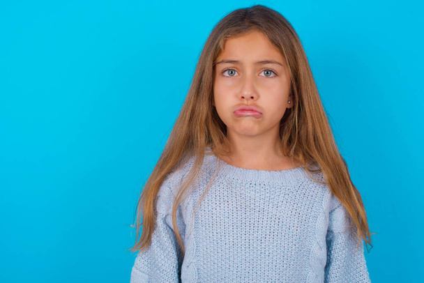 Displeased upset brunette kid girl wearing blue knitted sweater over blue background frowns face as going to cry, being discontent and unhappy as can't achieve goals,  Disappointed model has troubles - Photo, Image