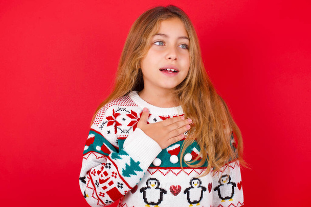 Joyful  beautiful girl wearing christmas sweater  expresses positive emotions recalls something funny keeps hand on chest and giggles happily. - Photo, Image
