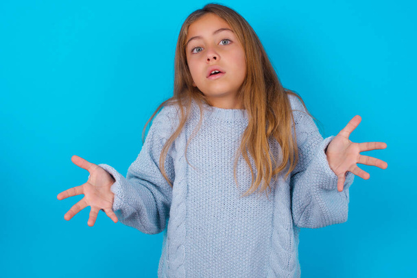 Frustrated brunetka dítě dívka na sobě modrý pletený svetr přes modré pozadí cítí zmatený a váhavý, pokrčí rameny v údivu, udržuje ústa široce otevřená, neví, co dělat. - Fotografie, Obrázek