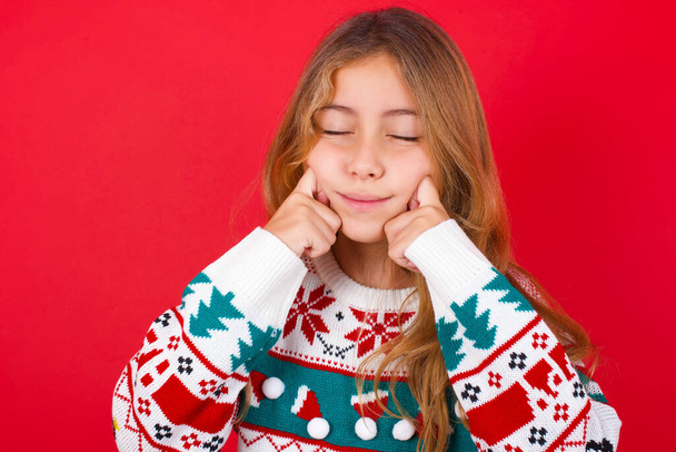 Tevreden mooi meisje dragen kerst trui met gesloten ogen houdt handen in de buurt wangen en glimlacht teder verbeeldt iets zeer aangenaam - Foto, afbeelding