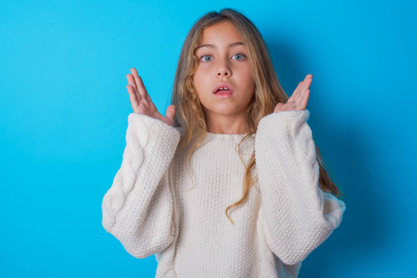 Surprised terrified brunette kid girl wearing white knitted sweater over blue background Gestures with uncertainty, stares at camera, puzzled as doesn't know answer on tricky question, People, body language, emotions concept - Foto, imagen
