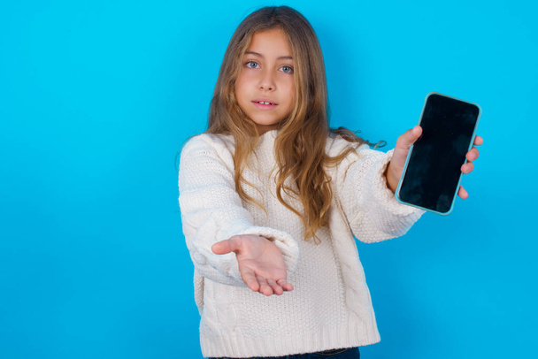 brunette lapsi tyttö yllään valkoinen neulotut villapaita sininen tausta mobiili. esittää älypuhelin. Mainoskäsite. - Valokuva, kuva