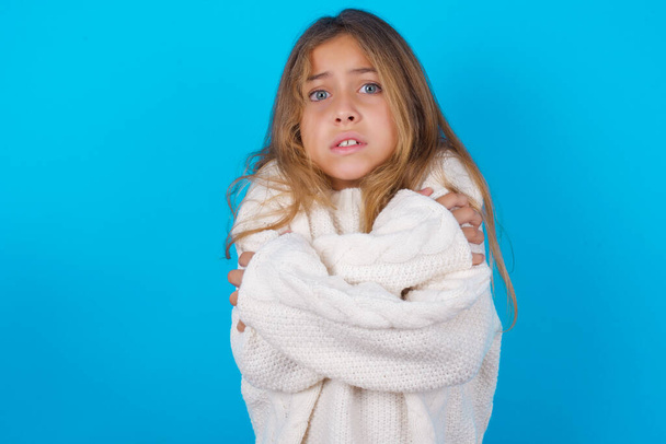 Desperate brunette kid girl wearing white knitted sweater over blue background trembles and feels cold, hugs oneself to warm up or feels scared notices something terrifying. - 写真・画像