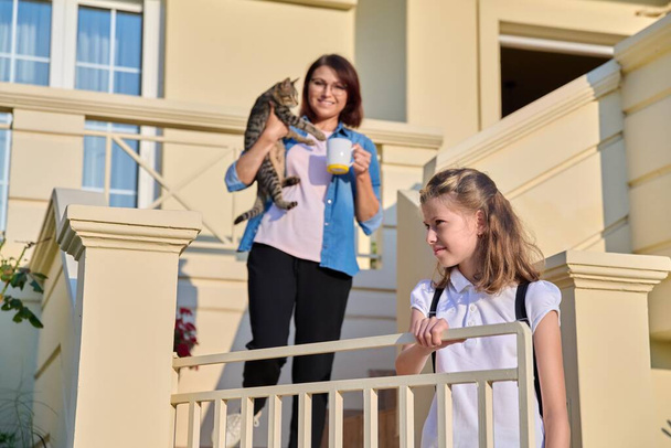 Mom withcat in arms on outdoor steps at home in morning with preteen daughter going to school - Foto, imagen