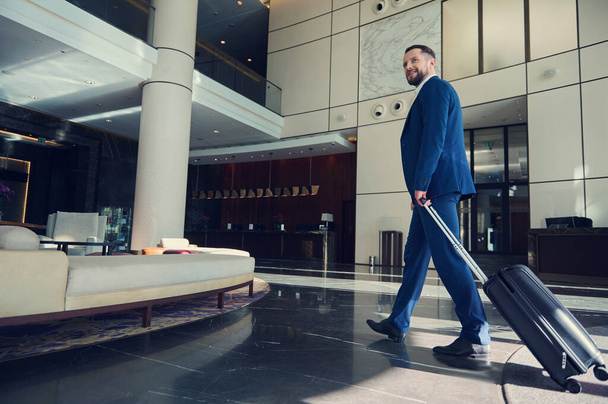 Przystojny mężczyzna, odnoszący sukcesy deweloper uśmiechnięty, spoglądający w bok, idący korytarzem, kierujący się z bagażem do salonu w nowoczesnym luksusowym budynku podczas podróży służbowej - Zdjęcie, obraz