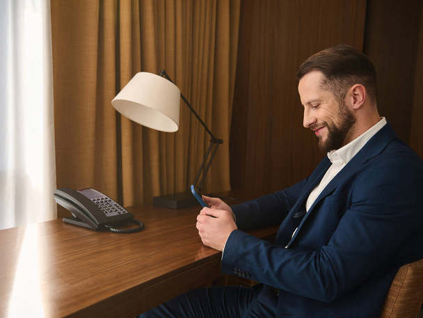 viaggiatore d'affari, uomo d'affari, imprenditore, investitore in tuta blu scuro a riposo in camera d'albergo, sorride mentre chatta sul cellulare durante il viaggio d'affari - Foto, immagini