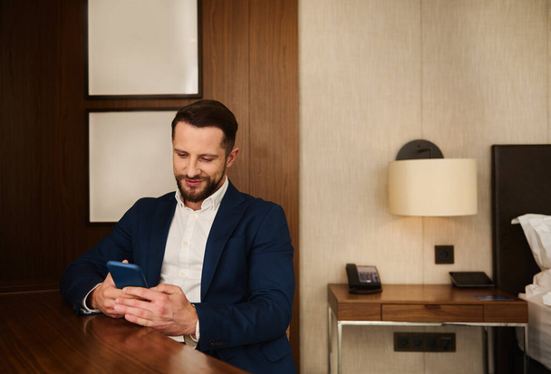 Confident portrait of a handsome prosperous business traveler, successful businessman, entrepreneur, investor in dark suit resting in hotel room, smiles while using mobile phone during business trip - Foto, Bild