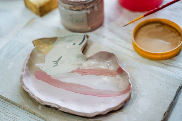 A plate of clay in the shape of a unicorn with a pink mane lies on the table, next to the paint and brush. Creativity in the pottery workshop. High quality photo - Φωτογραφία, εικόνα