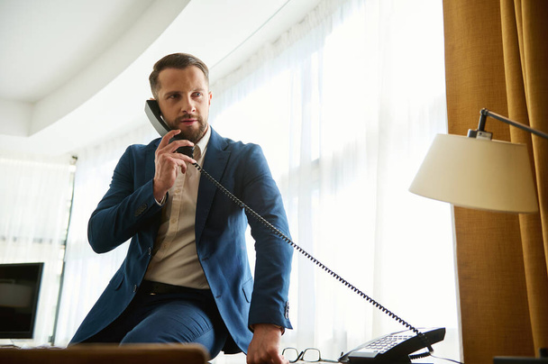 Hombre de negocios guapo y seguro de sí mismo, hombre atractivo, inversor financiero próspero en un viaje de negocios, sentado en la esquina de la mesa de la oficina en la habitación del hotel y llamando a la recepción por teléfono fijo - Foto, imagen