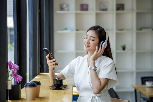  asiatique femme d'affaires assis près de la fenêtre en verre et en streaming écouter de la musique du téléphone mobile au casque sans fil et boire du café heureux   - Photo, image